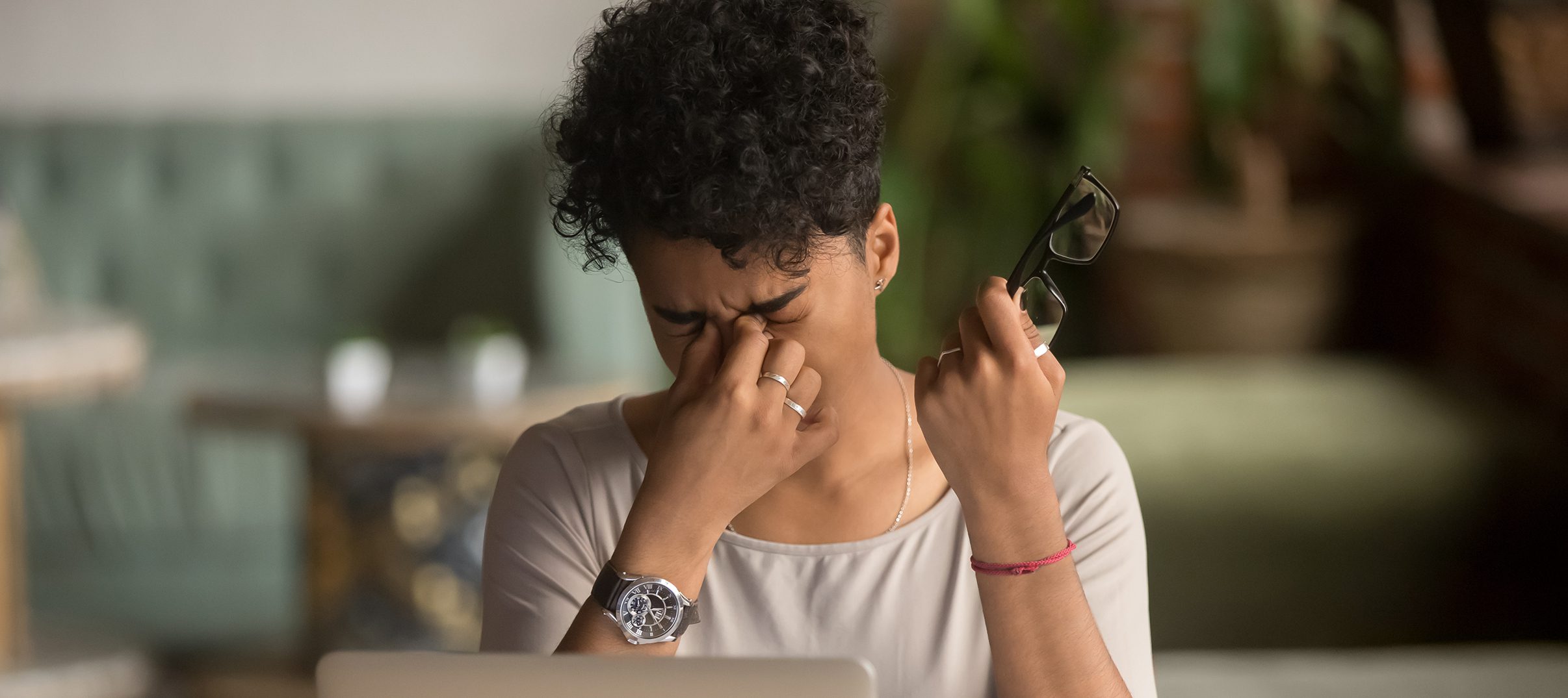A person sits at a table, holding their glasses from une lunetterie à Montréal in one hand and pinching the bridge of their nose with the other, appearing stressed while looking at a laptop.
