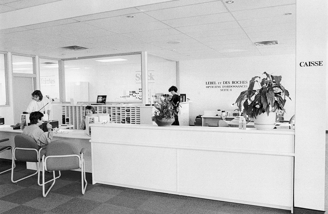 Black and white photo of a bank or office reception area, with several people working at desks and counters. A large plant sits on the front counter, and CAISSE is written on the wall to the right—Vu à la lunetterie à Montréal.