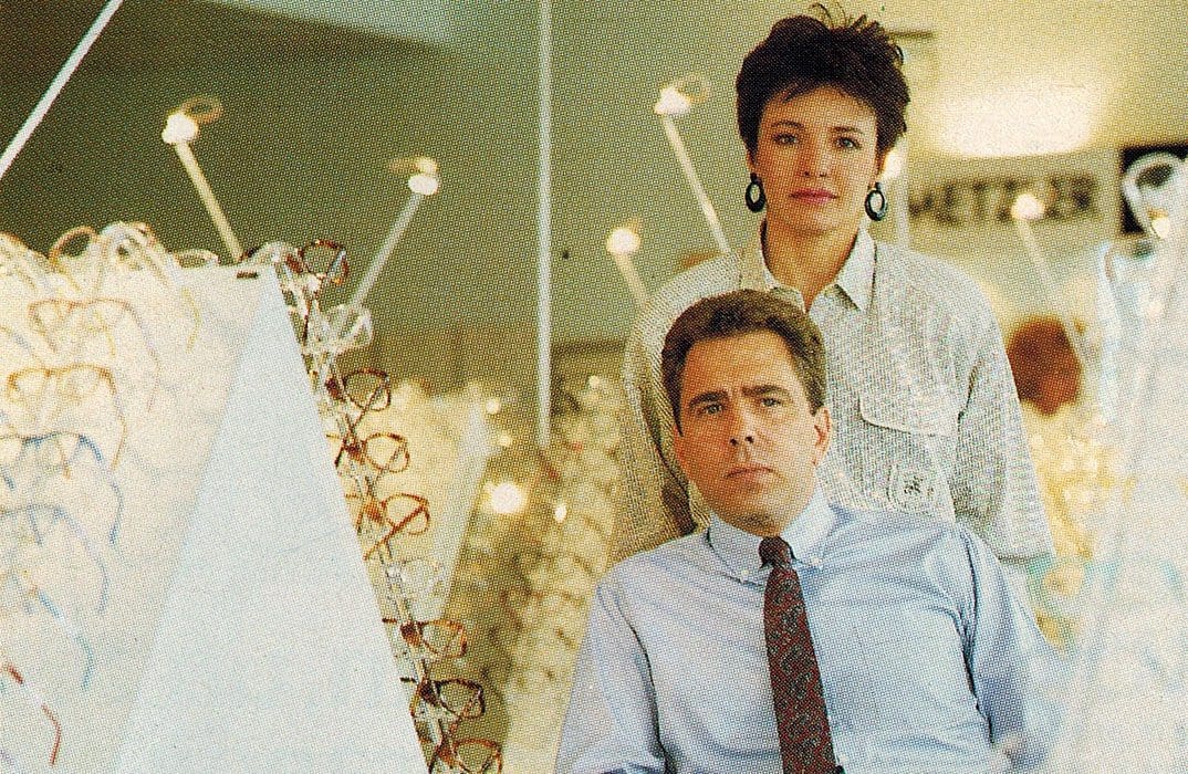 A man in a shirt and tie sits in front of a woman in a patterned blouse, both surrounded by displays of eyeglasses at Vu, la lunetterie à Montréal avec le plus grand inventaire de montures à Montréal, in a brightly lit optical store.