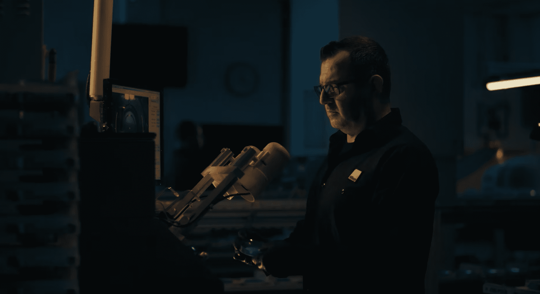 A man in a dark room at Vu, le plus grand inventaire de montures à Montréal, works at his desk surrounded by computer screens and equipment. He wears glasses and a dark shirt, focusing intently on his task under dim, bluish lighting.