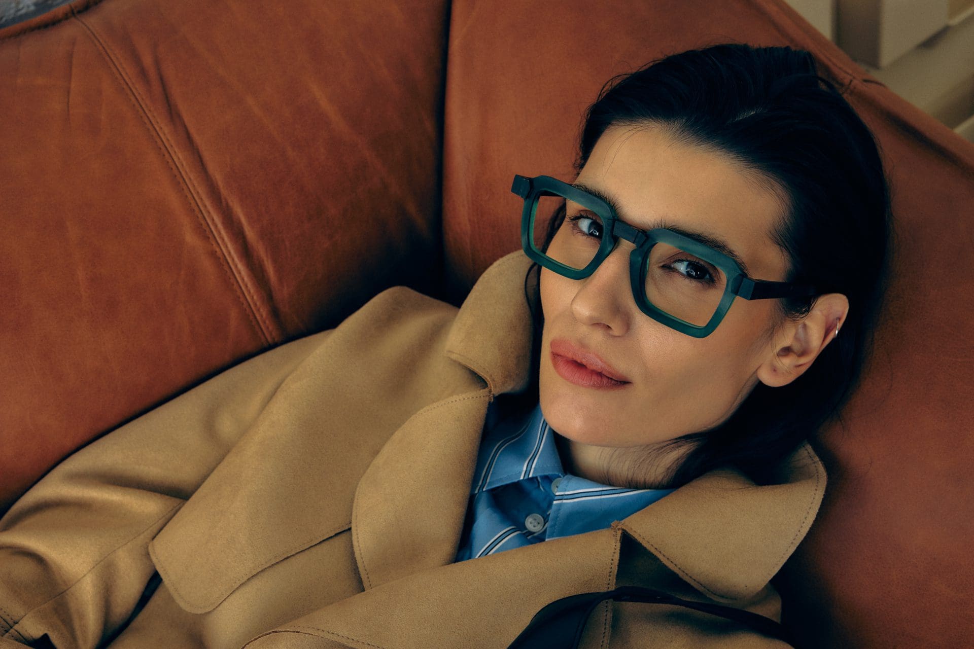 A woman with dark hair, wearing statement geometric glasses from Vu, reclines on a brown leather couch in a tan coat and blue collared shirt, looking directly at the camera. Découvrez le plus grand inventaire de montures à Montréal.