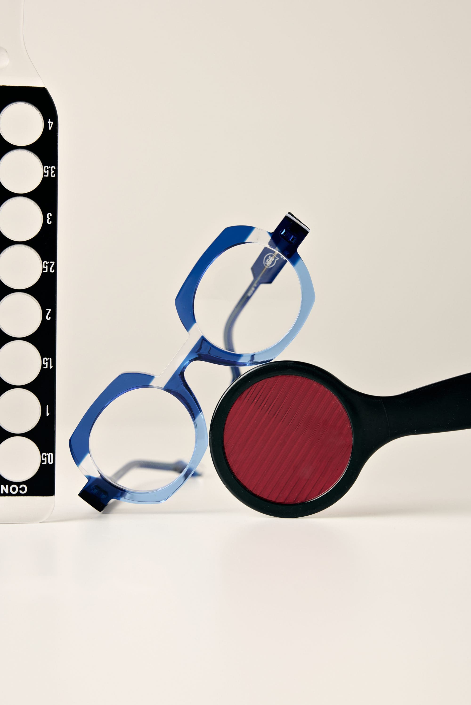 Blue eyeglasses rest on a white surface next to a black-and-white vision test chart, with a red lens tool placed in front—discover Vu, la lunetterie à Montréal avec le plus grand inventaire de montures à Montréal.