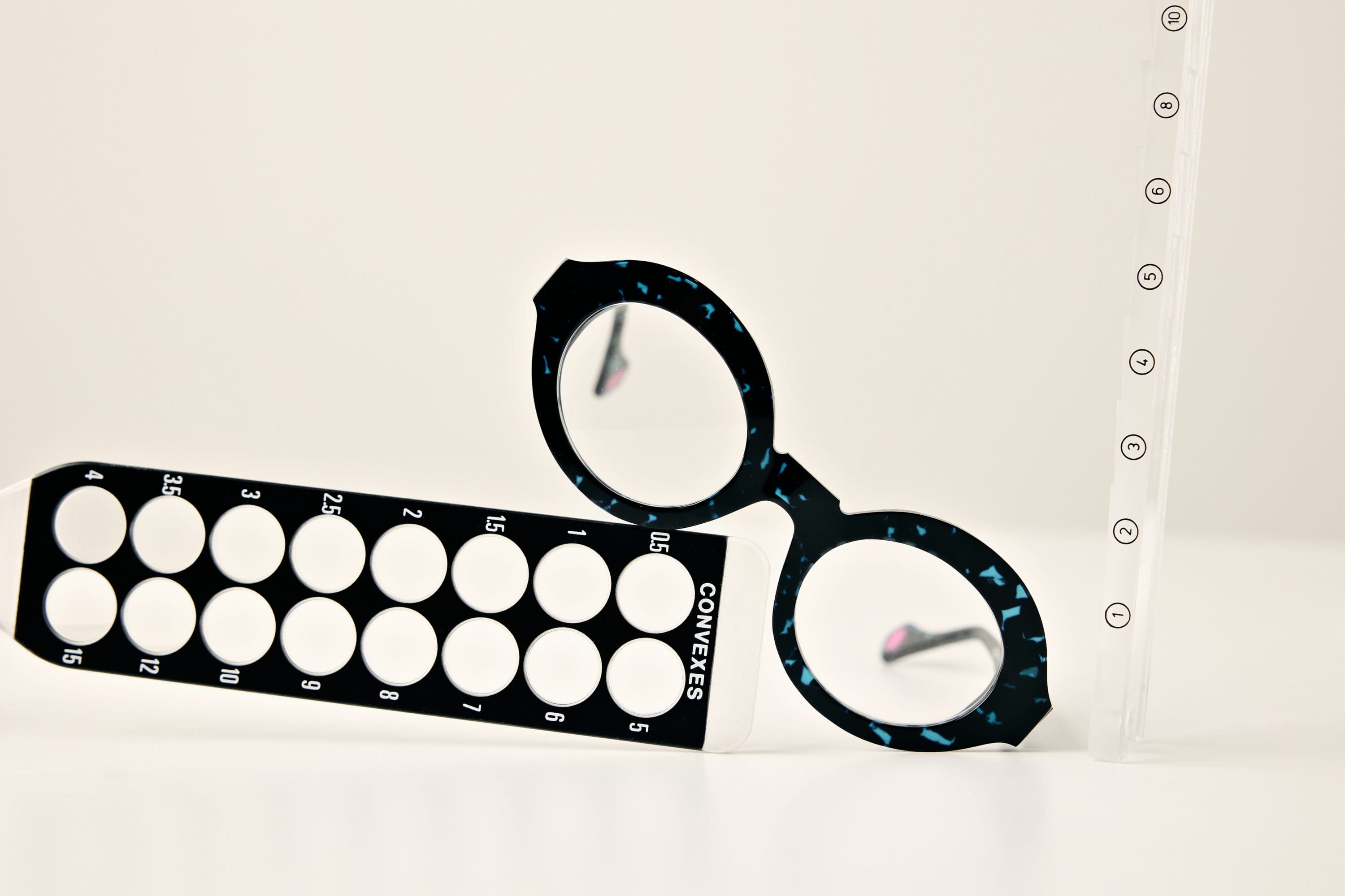 Round, black-rimmed glasses with blue specks rest between a black vision test tool and a clear vertical ruler, all set against a plain white background—showcasing Vu: le plus grand inventaire de montures à Montréal.