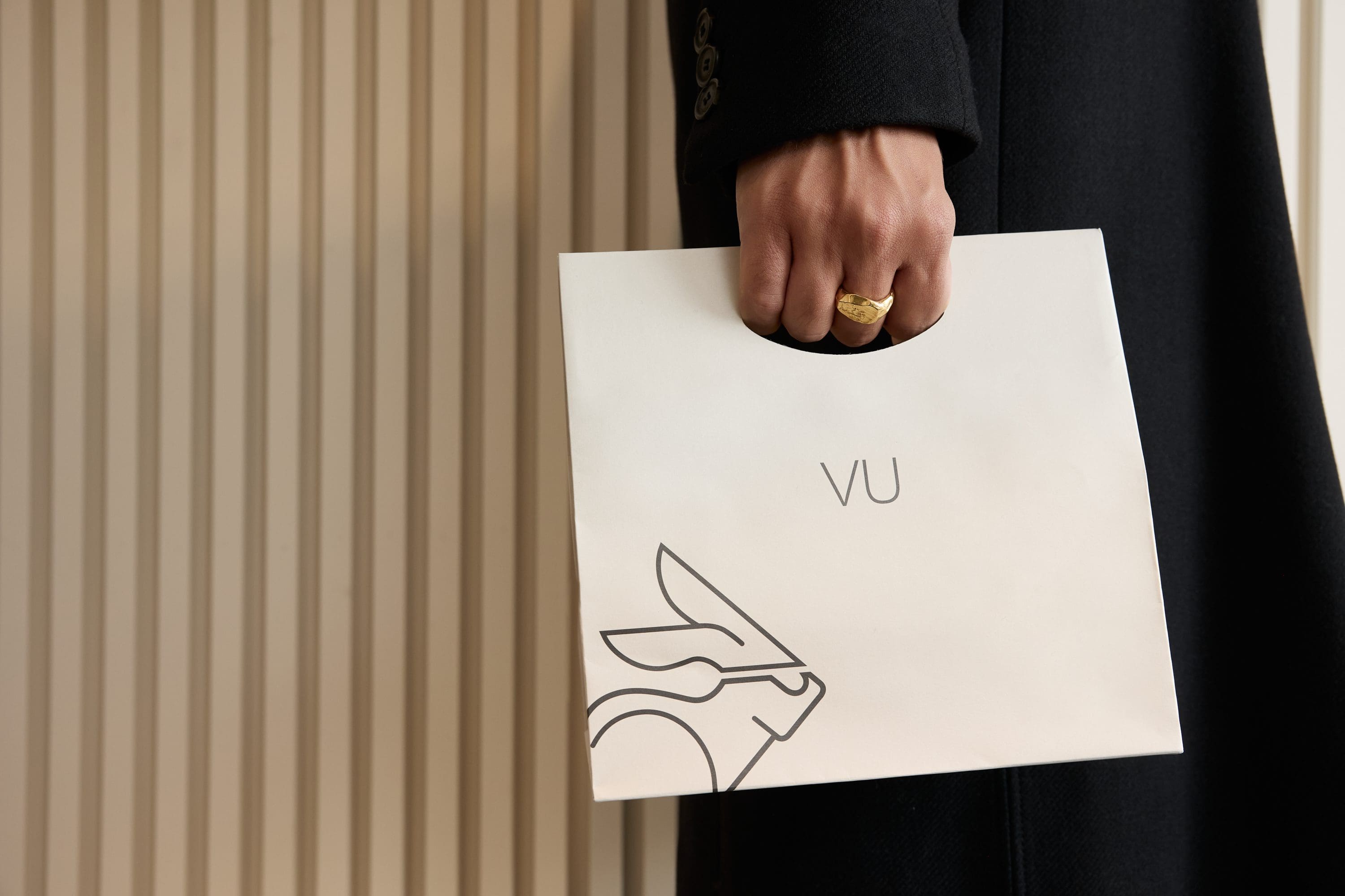 A person wearing a black coat and gold ring holds a white shopping bag with a minimalist animal illustration and the letters Vu, representing lunetterie à Montréal, against a background of vertical beige panels.