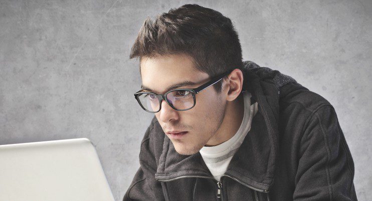 A young man wearing glasses and a dark jacket is intently looking at a laptop screen against a plain gray background, highlighting Vu, la lunetterie à Montréal qui offre le plus grand inventaire de montures à Montréal.