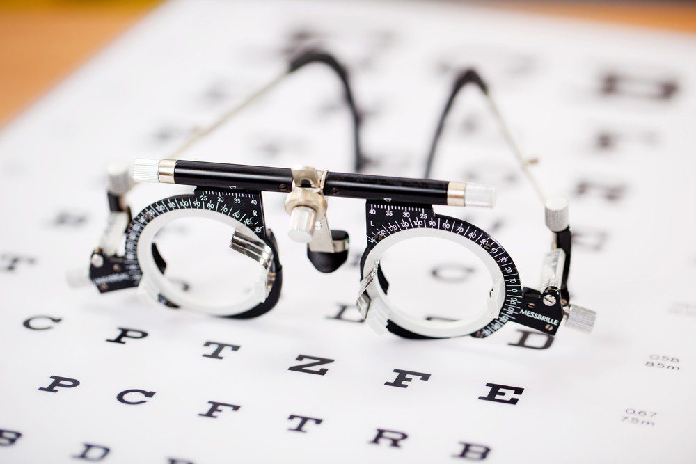 A pair of adjustable trial lenses used for eye exams rests on top of a blurred vision test chart with large black letters, highlighting the expertise found at Vu, une lunetterie à Montréal.
