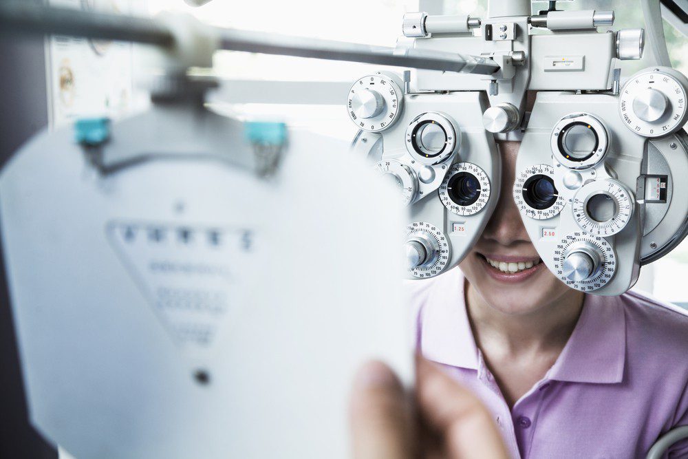 A person smiling while undergoing an eye exam at Vu, une lunetterie à Montréal, looking through a phoropter as an optometrist holds up a vision test chart.