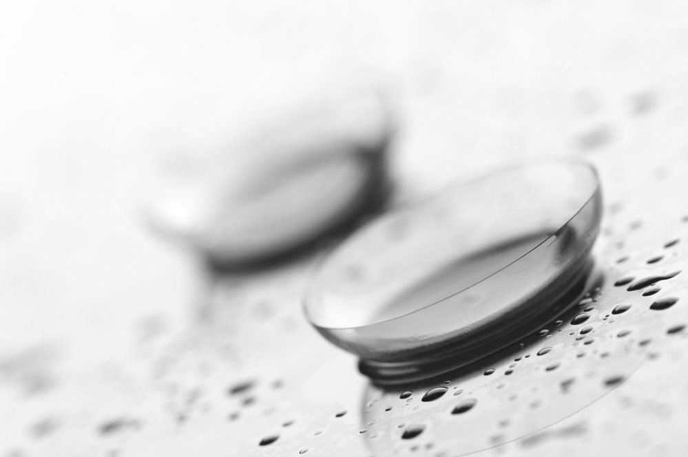Two transparent contact lenses lie on a wet surface with water droplets around them, photographed in black and white—a subtle nod to lunetterie à Montréal and its dedication to clear vision.