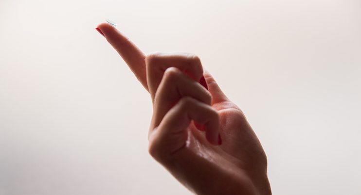 A close-up of a hand with a raised index finger, holding a contact lens from Vu on the fingertip against a light, blurred background—perfect for those seeking lunetterie à Montréal.