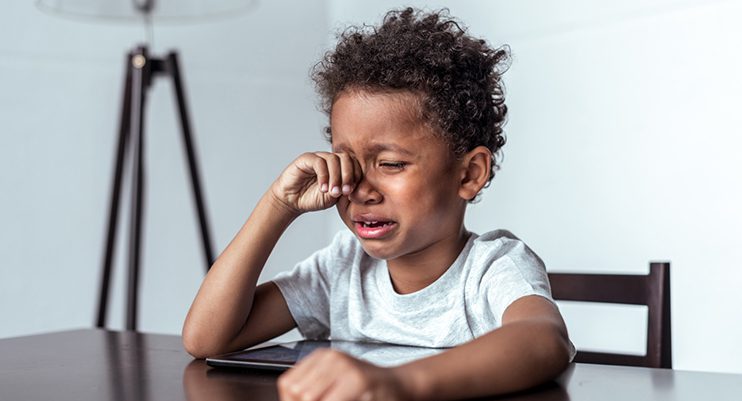 A young child with curly hair sits at a table, crying and rubbing their eyes with one hand, while a tablet lies on the table—perhaps in need of Vu, le plus grand inventaire de montures à Montréal, to help ease eye strain.