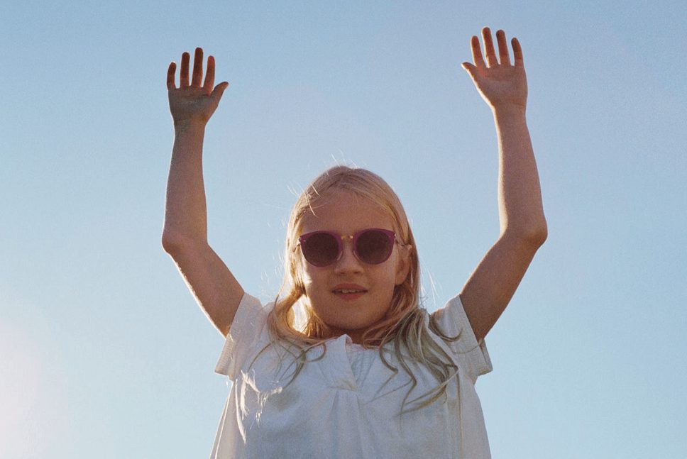 A young girl with long blonde hair wears Vu sunglasses and a white shirt, smiling with both arms raised high against a clear blue sky.