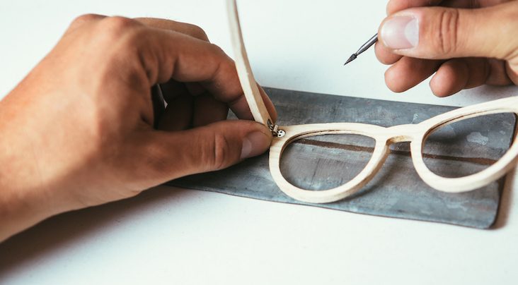 A person uses a small screwdriver to repair the hinge of light-colored eyeglass frames, which rest on a cloth—showcasing Vu, le plus grand inventaire de montures à Montréal, votre référence en lunetterie à Montréal.