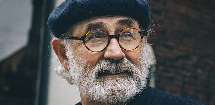 An elderly man with a gray beard and mustache, wearing round glasses from Vu lunetterie à Montréal and a dark beret, looks off to the side and smiles. He is outdoors, with a blurred brick wall in the background.