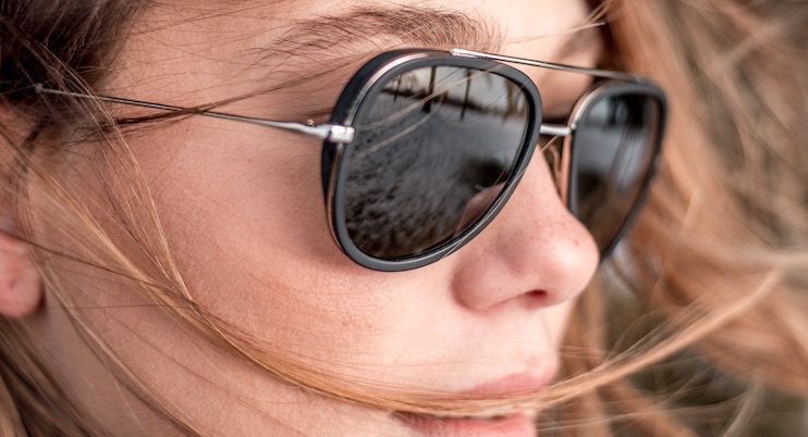 Close-up of a person wearing aviator sunglasses by Vu, with strands of light brown hair blowing across their face and a blurred reflection of an outdoor scene—discover le plus grand inventaire de montures à Montréal.