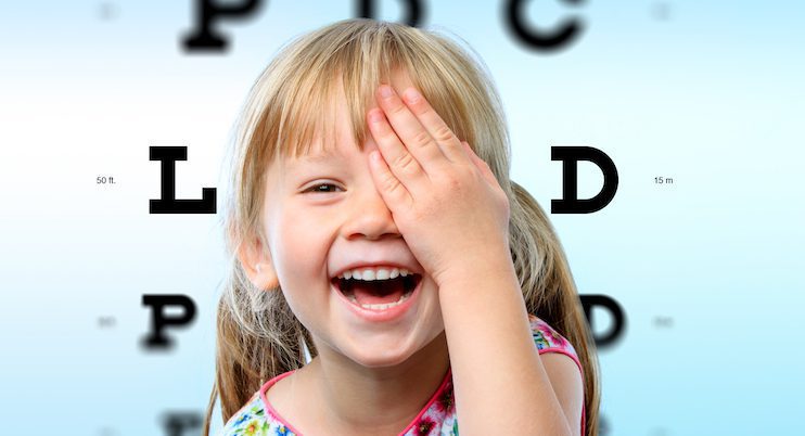 A smiling young girl covers one eye with her hand during an eye exam at Vu, une lunetterie à Montréal offrant le plus grand inventaire de montures, standing in front of a blurry vision chart with large letters.