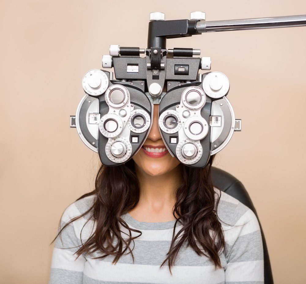 A woman with long brown hair and a striped shirt smiles while sitting behind a phoropter during an eye exam at Vu, la lunetterie à Montréal.