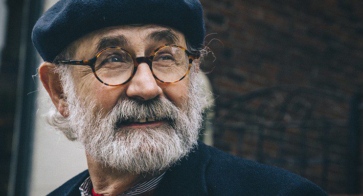 An older man with a white beard and mustache, wearing round glasses from le plus grand inventaire de montures à Montréal, a dark beret, and coat, looks off to the side and smiles softly. A blurred brick wall is in the background.
