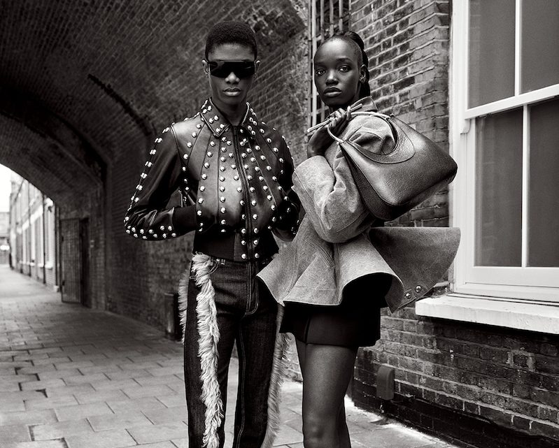 Two stylish models pose on a city street. One wears Vu lunetterie à Montréal sunglasses, a studded jacket, and fur-trimmed pants; the other, with a large bag, sports an oversized jacket and skirt. The monochrome scene is chic and fashion-forward.