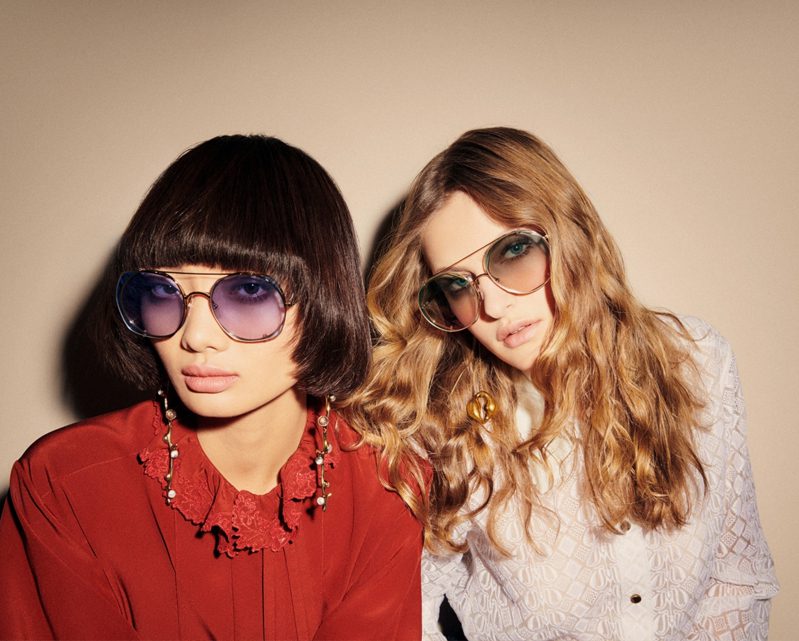 Two women pose together wearing large, tinted sunglasses from Vu, la lunetterie à Montréal. One has a short dark bob and a red blouse with statement earrings; the other sports long blonde waves and a white textured top.