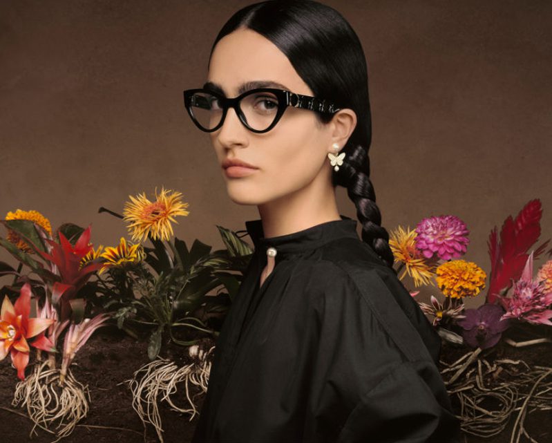 A woman with long dark hair in a braid, wearing black glasses from Vu, a renowned lunetterie à Montréal, and pearl earrings, poses before colorful flowers and exposed roots against a rich brown background.