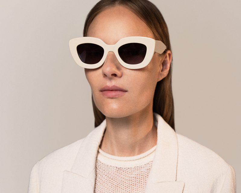 A woman with straight brown hair wears white cat-eye sunglasses from Vu, a white textured top, and a light-colored blazer, posing against a neutral background—showcasing le plus grand inventaire de montures à Montréal.