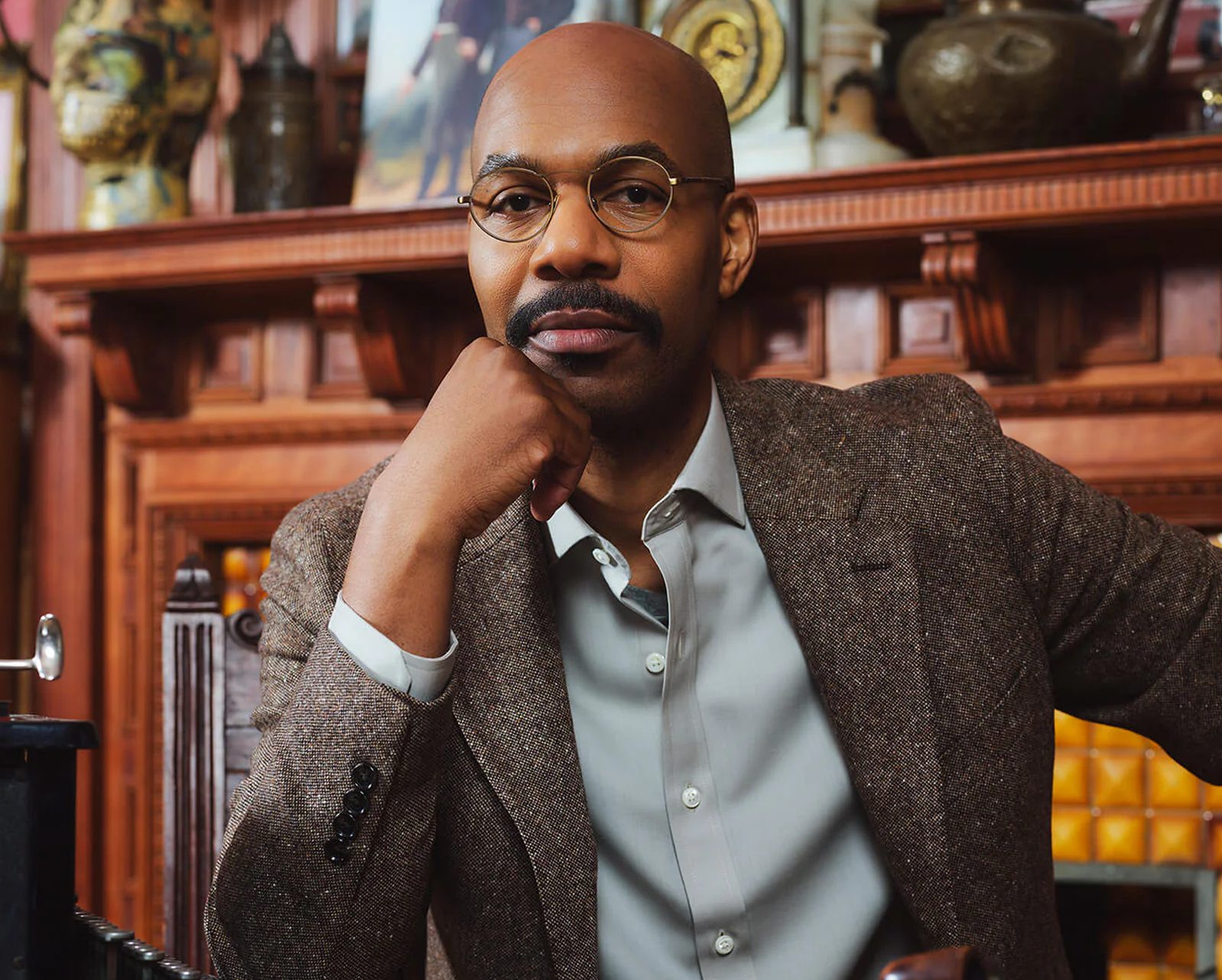 A man with glasses from la lunetterie à Montréal, wearing a brown tweed blazer and gray shirt, sits with his hand on his chin in an ornate room filled with wooden shelves and decorative objects.