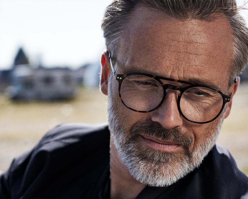 A middle-aged man with gray hair and a beard wears black-rimmed glasses from a lunetterie à Montréal, looking thoughtfully downward. The blurred background hints at sunlight and an outdoor setting.