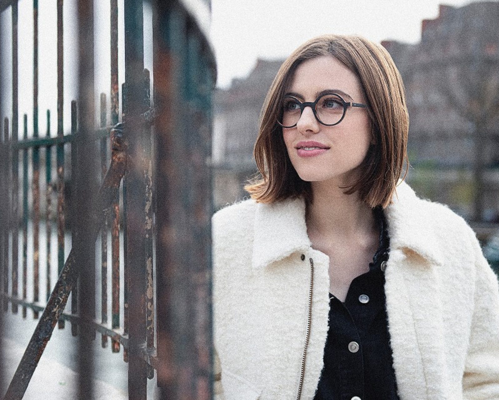 A woman with shoulder-length brown hair and round glasses, wearing a white fuzzy coat over a black top, stands outdoors near a metal fence, looking to the side and smiling softly—capturing the chic style of lunetterie à Montréal.