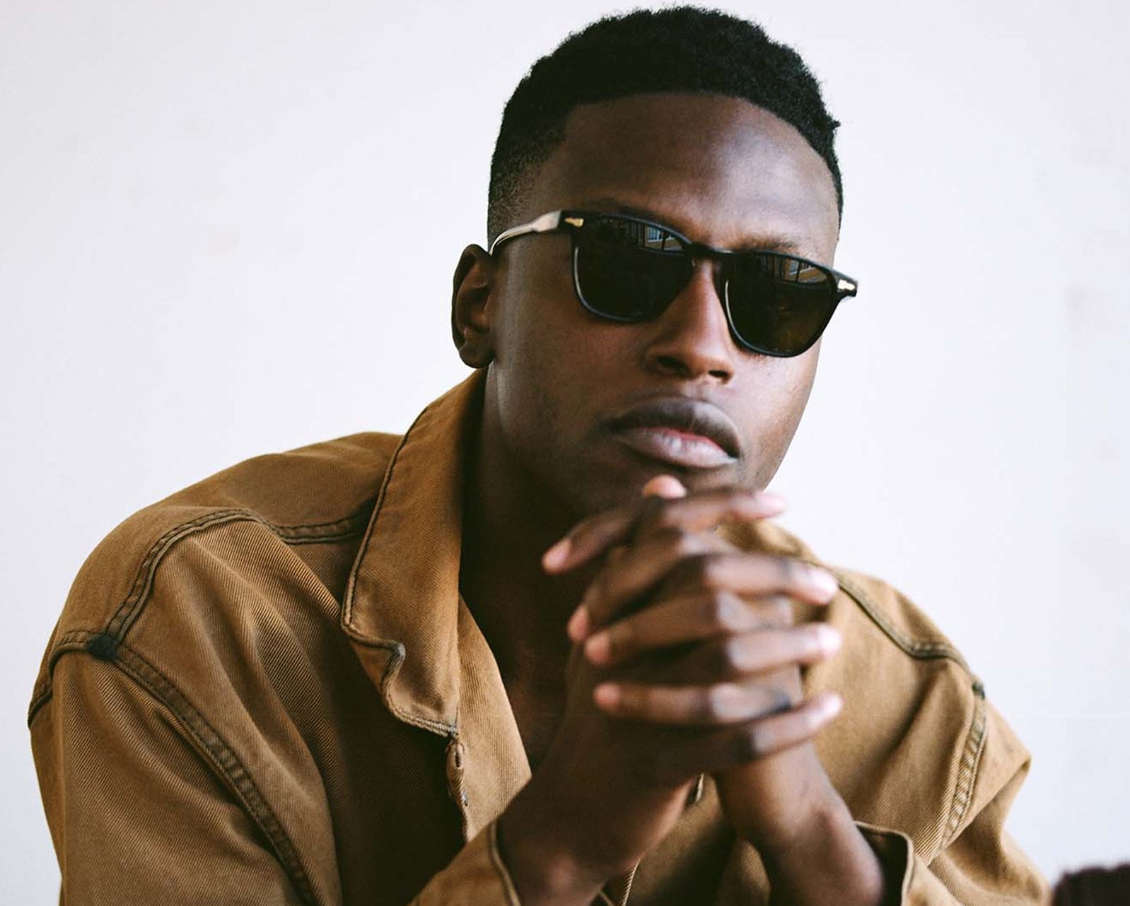 A person wearing dark sunglasses from Vu, the lunetterie à Montréal with le plus grand inventaire de montures, sits with their hands clasped, looking directly at the camera against a plain light background.