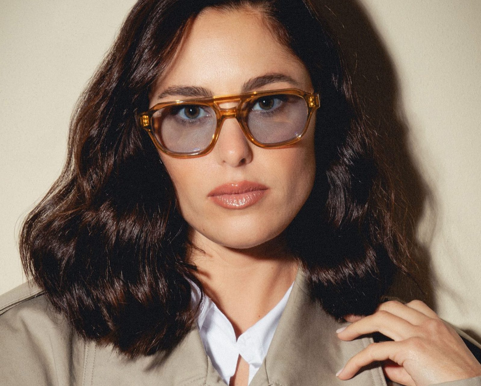 A woman with wavy dark brown hair wears large, amber-colored Vu sunglasses and a beige coat over a white shirt, looking directly at the camera—a chic look from la plus grande lunetterie à Montréal.