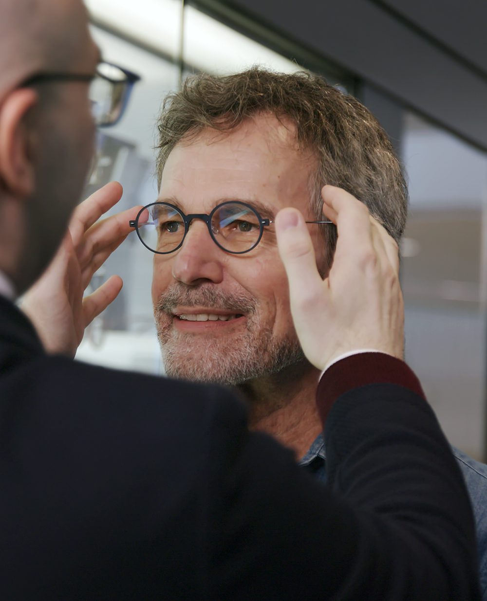 An older man with salt-and-pepper hair and a beard smiles as another person helps him adjust round eyeglasses on his face at Vu, la lunetterie à Montréal ayant le plus grand inventaire de montures à Montréal.